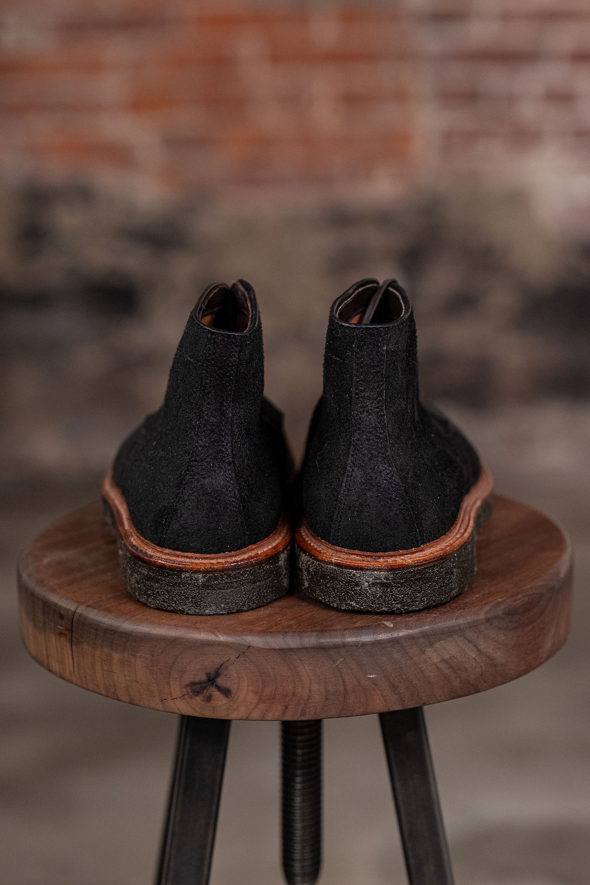 Pair of Alden Plain Toe boots on a wooden stool with a blurred brick wall background - back view