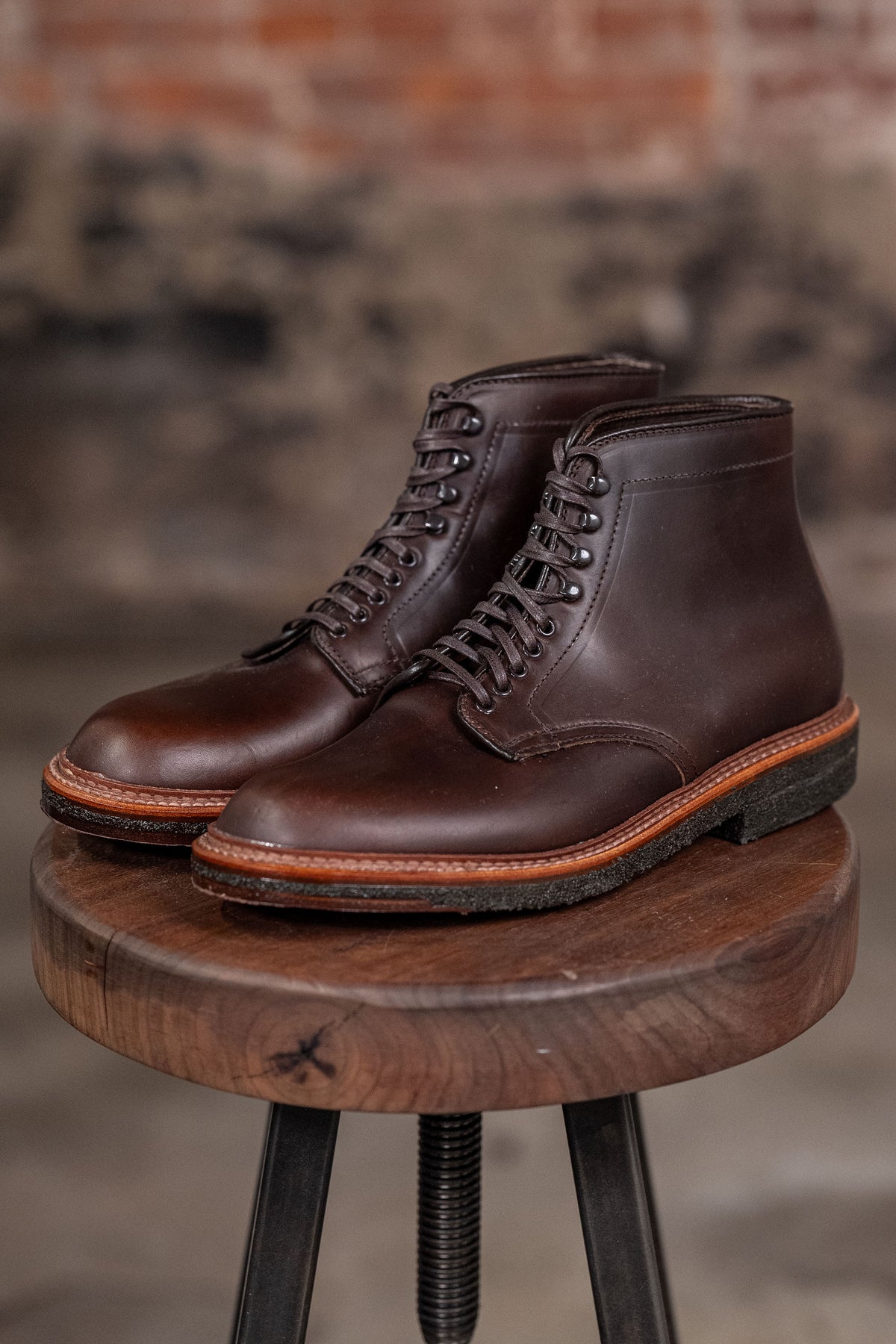Pair of Alden Plain Toe boots on a wooden stool with a blurred brick wall background - three quarter view