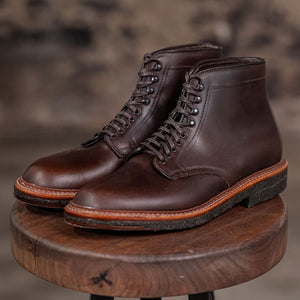 Pair of Alden Plain Toe boots on a wooden stool with a blurred brick wall background - three quarter view