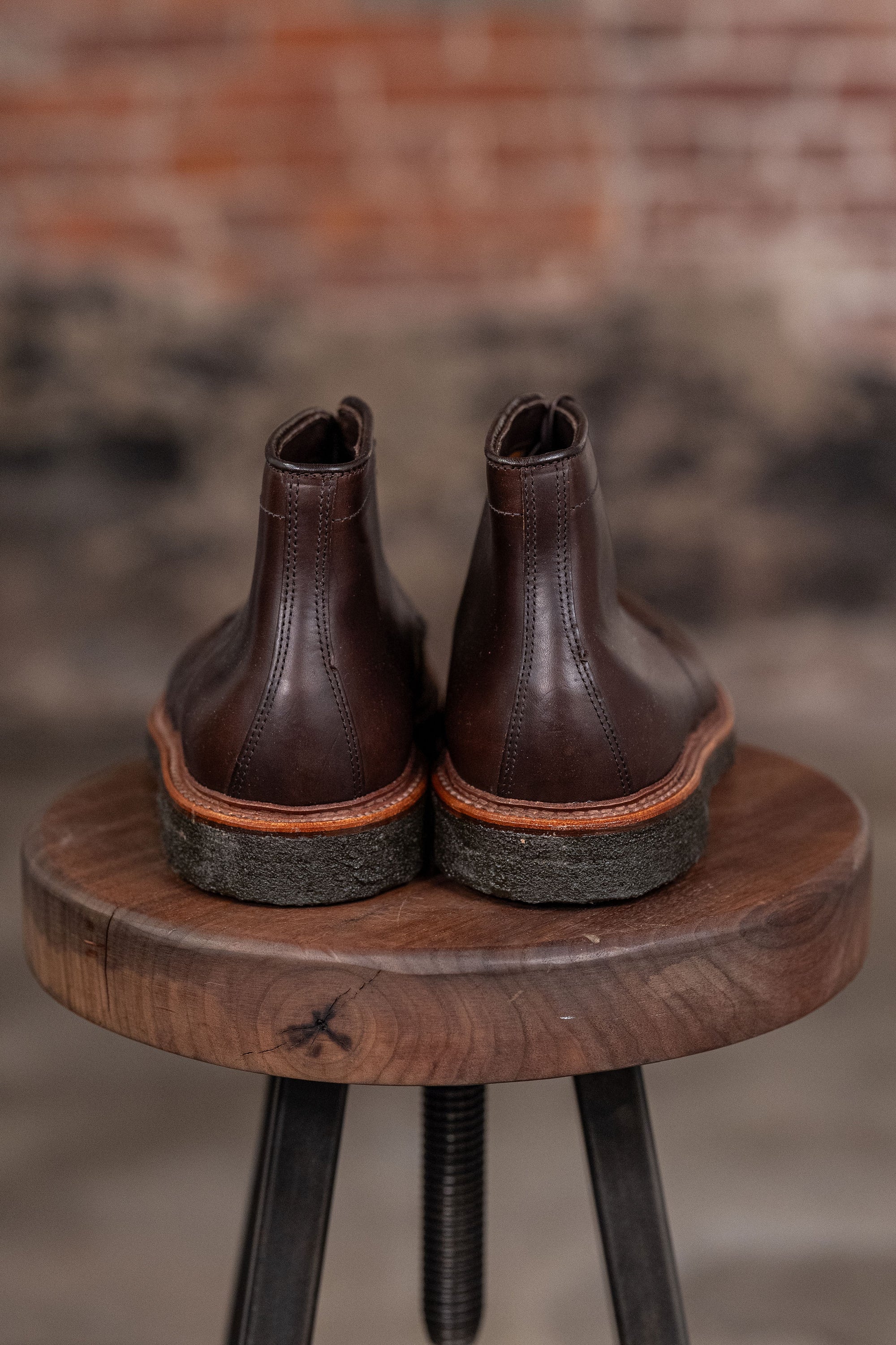 Pair of Alden Plain Toe boots on a wooden stool with a blurred brick wall background - back view