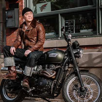 Franklin and Poe founder wears a leather jacket and denim while sitting on a Triumph motocycle in front of the old shop window